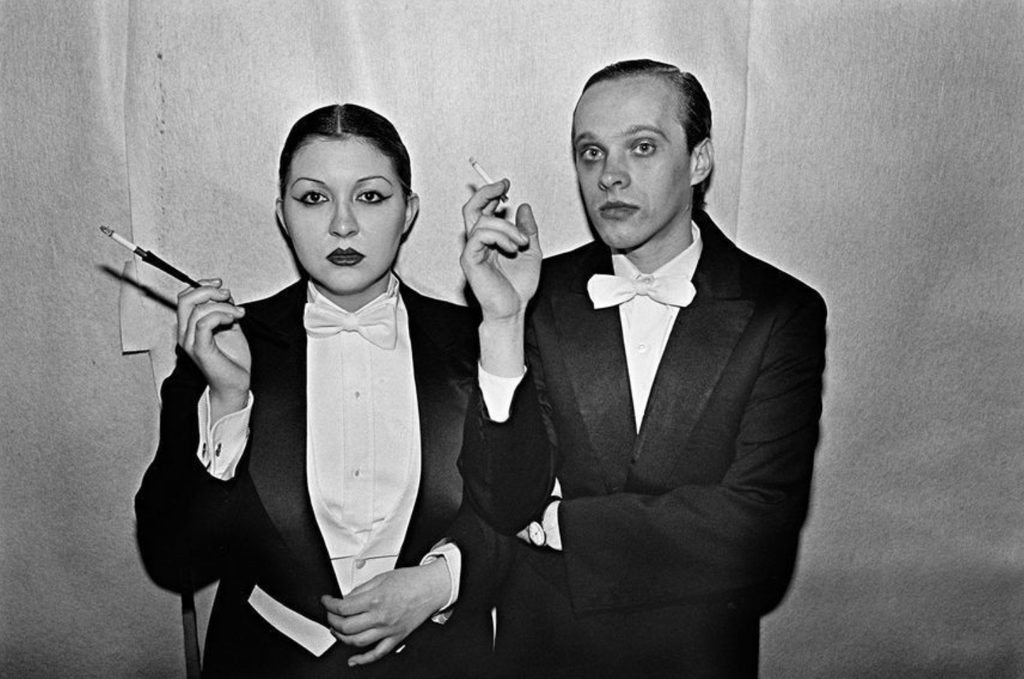 A couple dressed in matching tuxedos with cigarettes, standing in front of a curtain.