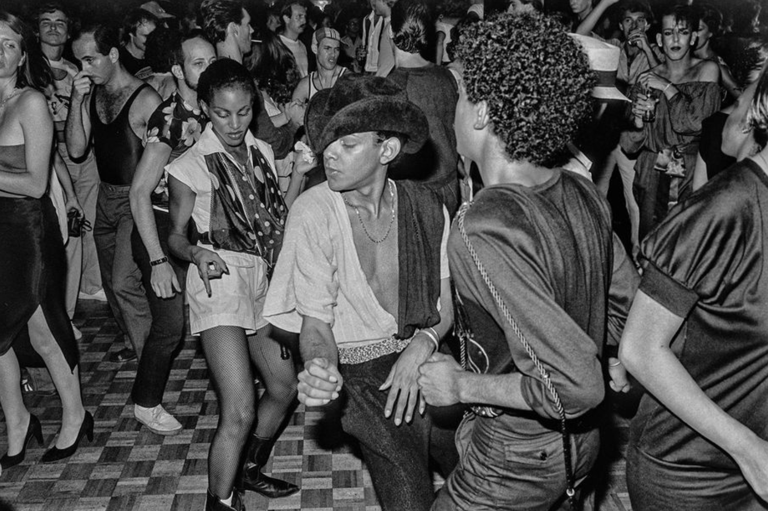 A crowded nightclub dance floor with dancers gathered around a central figure wearing a hat.