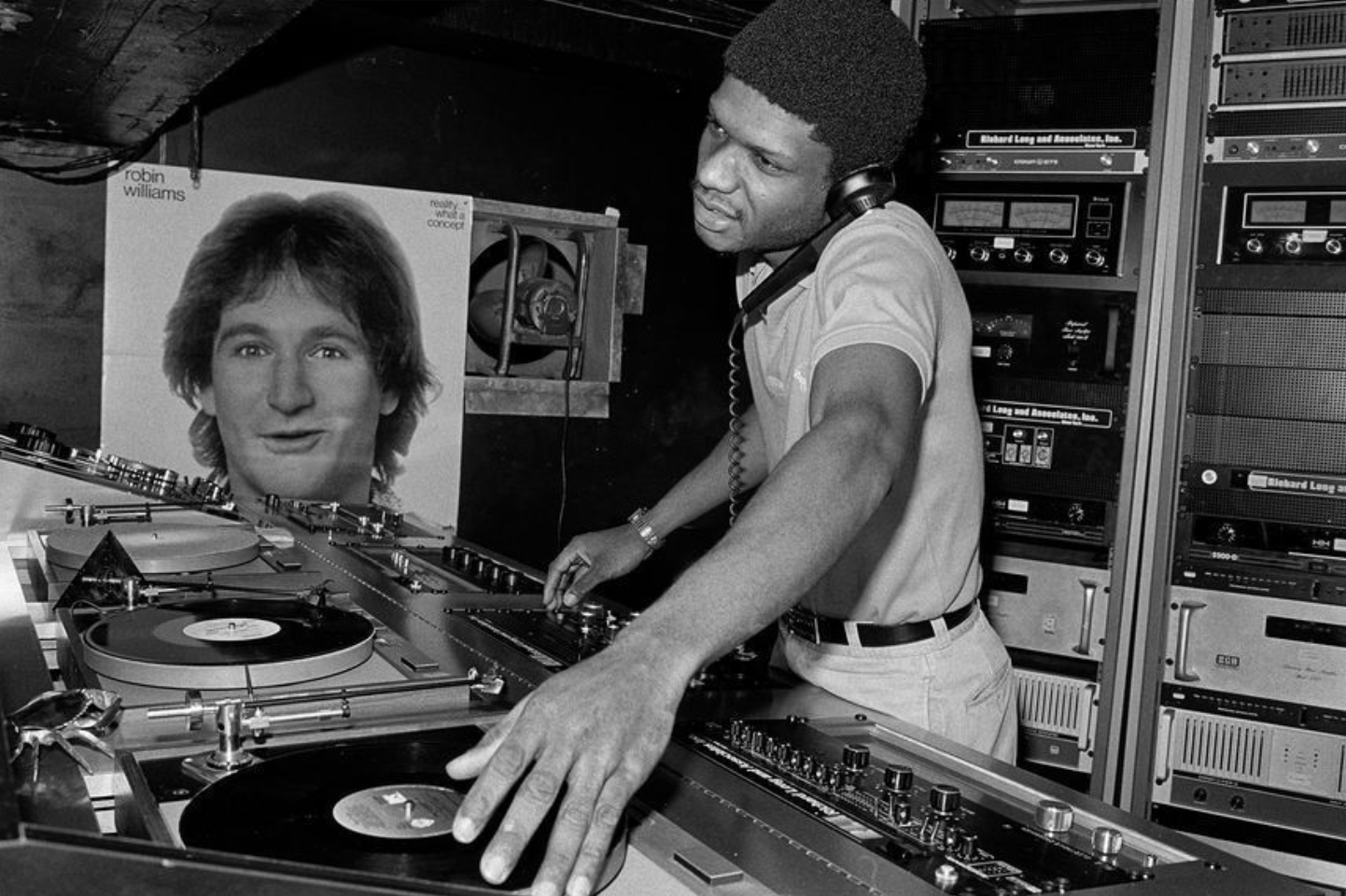 A DJ at work behind turntables in a nightclub booth, surrounded by amplifiers and sound equipment.