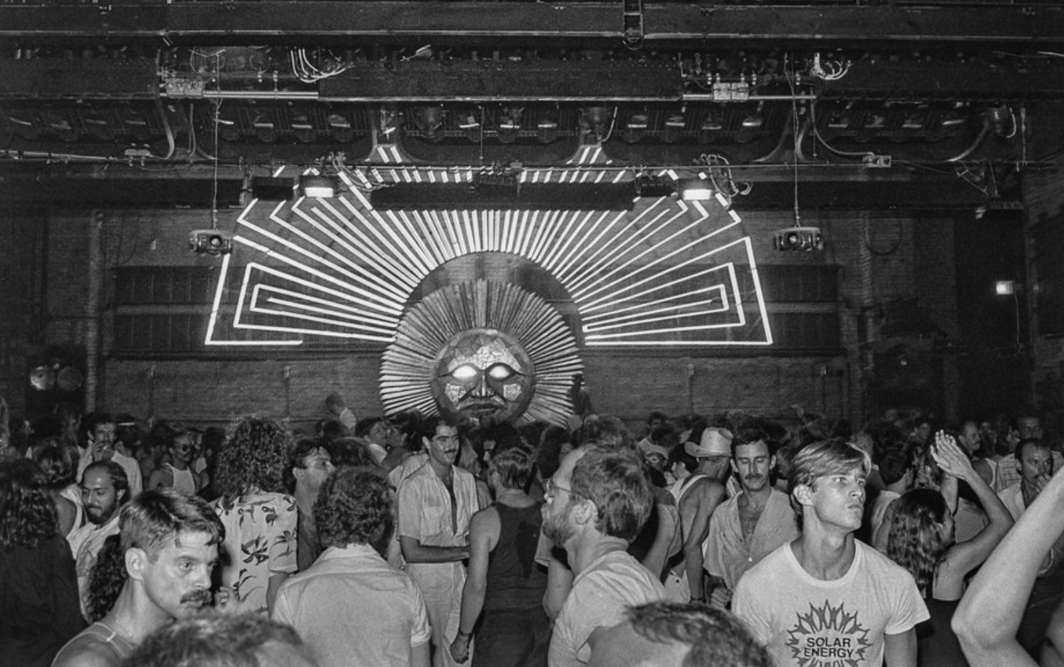 Wide view of a packed New York nightclub with a large geometric neon light installation above the crowd.